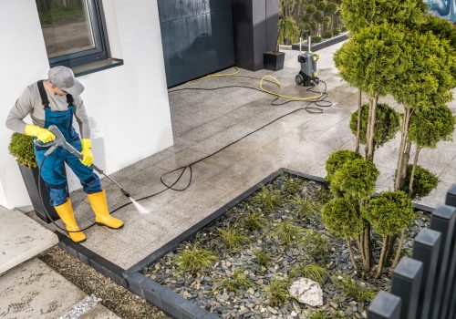 Caucasian Men Pressure Washing House Surrounding Concrete Elements. Cleaning Paths and Driveway Using Powerful Washer.