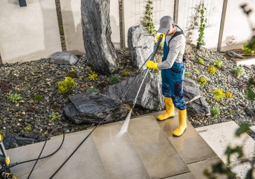 Caucasian Man in Protective Work Wear Cleaning Garden Paths with Pressure Washer. Decorative Stone and Gravel Aggregates in the Background. Spring Season.