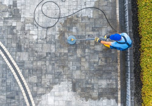 A worker uses a pressure washer to clean a stone patio surrounded by green foliage on a clear day, creating a refreshed outdoor space.