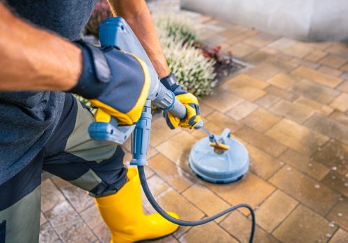 A person in yellow boots and gloves operates a pressure washer on a stone patio, effectively cleaning the surface in a residential backyard setting.