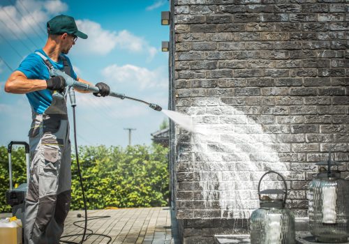 Brick House Wall Pressure Washing with Special Cleaning Detergent. Caucasian Men in His 30s. Taking Care of the Building Elevation.