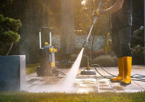 A worker in yellow boots uses a pressure washer to clean a patio area under bright sunlight, showcasing effective outdoor maintenance.