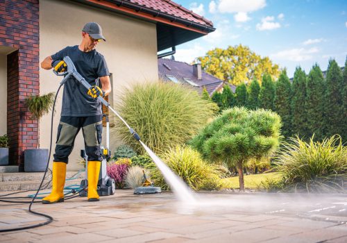 A man is using a power washer to clean the patio in a beautiful garden filled with greenery and sunshine.